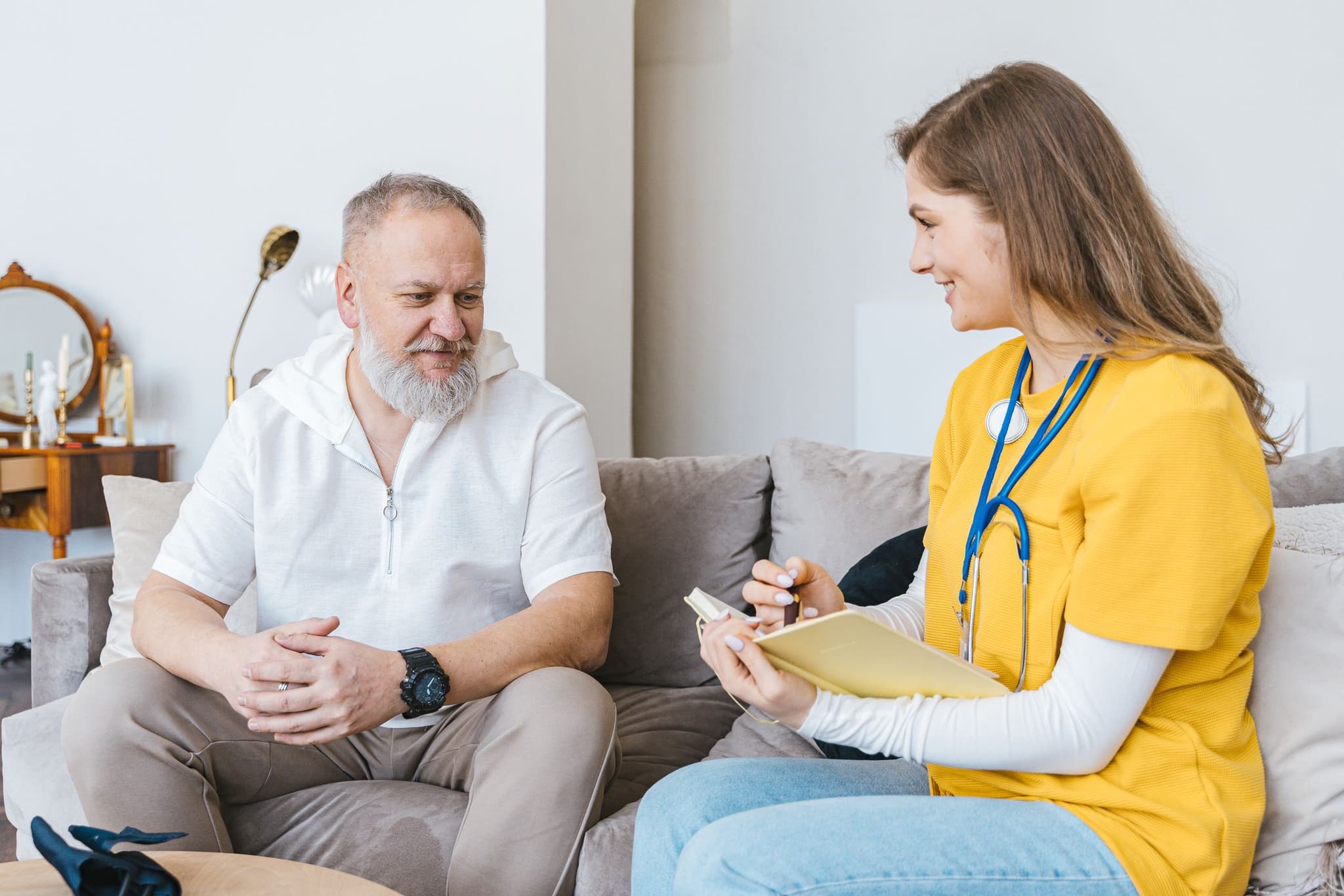 Pflegefachfrau mit Stethoskop und Notizbuch im Gespräch mit einem älteren Klienten auf dem Sofa zuhause